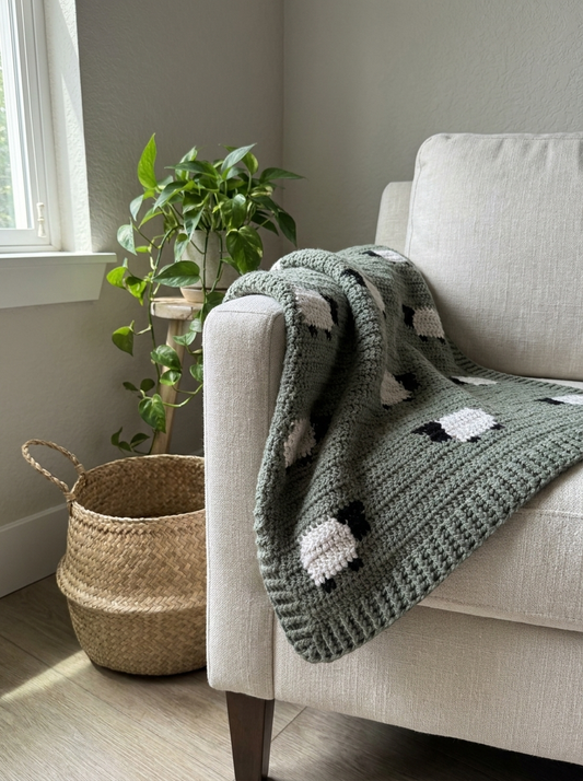 Verdant Glen Crochet Baby Blanket Pattern - sage green crochet blanket with sheep motifs folded over a linen armchair
